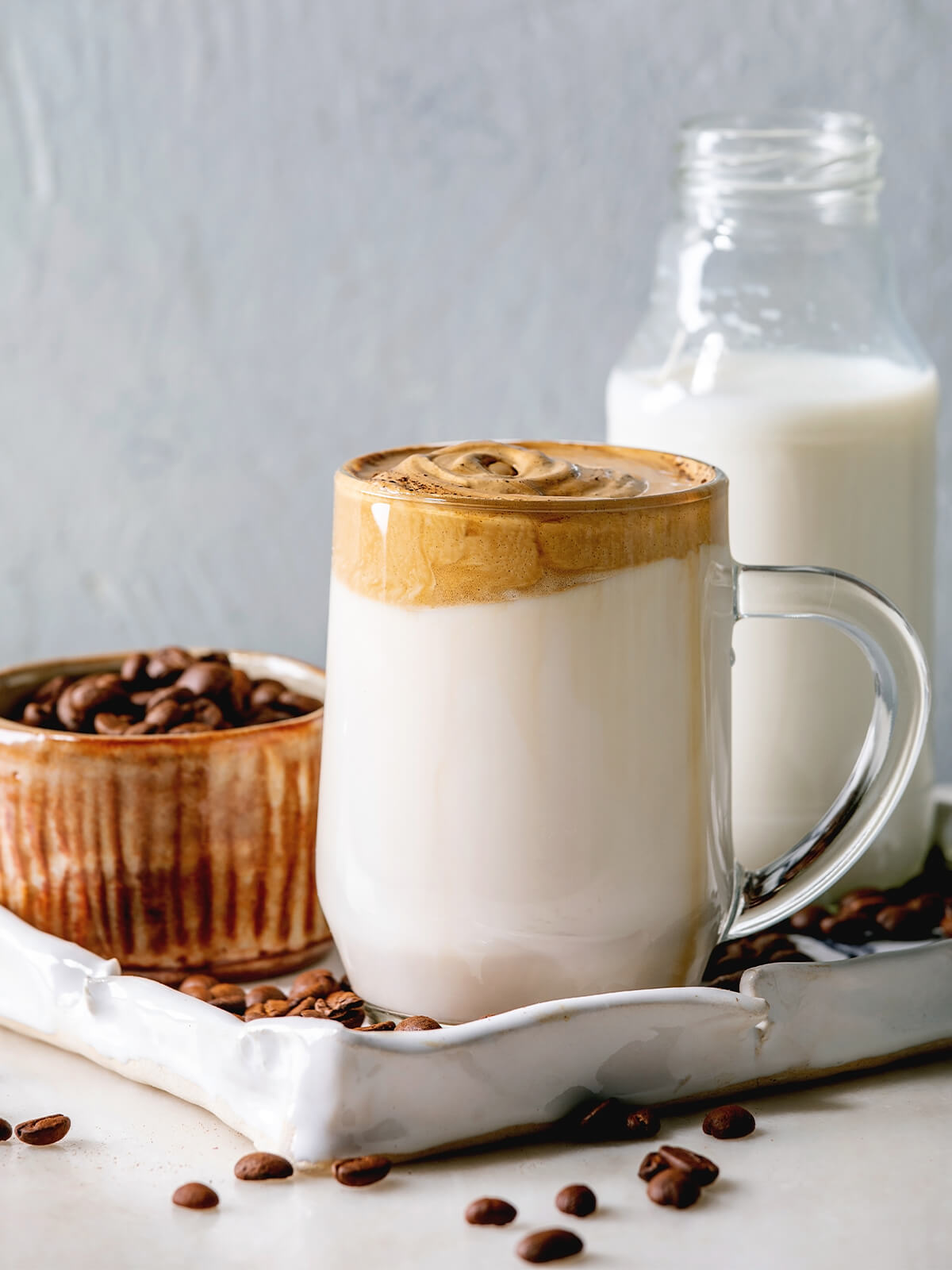 Dalgona-Kaffee im Glasbecher auf weißer Schale, daneben eine Schüssel Kaffeebohnen und eine Flasche Milch