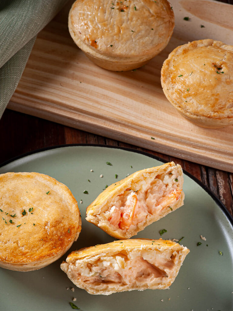 Goldgelbe Empadas de Camarao von oben fotografiert, im Vordergrund eine aufgeschnittene und eine ganze Empada auf einem grünen Teller mit Anschnitt der cremigen Füllung, dahinter weitere ganze Pastetchen auf einem Holzbrett