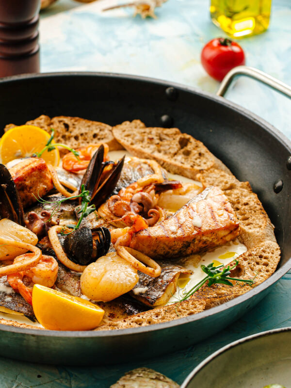 Fisch- und Meeresfrüchte-Pfanne in Sahnesauce mit gegrilltem Lachs, weißem Fisch, Garnelen, Calamari, Miesmuscheln, Oktopus und Jakobsmuscheln, dazu Zitrone, Thymian und geröstetes Brot