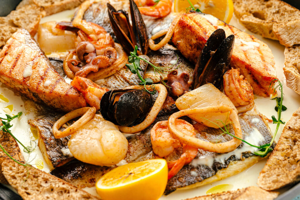 Nahaufnahme einer Fisch- und Meeresfrüchte-Pfanne in heller Sahnesauce mit gegrilltem Lachs, weißem Fisch, Garnelen, Calamari, Miesmuscheln, Oktopus und Jakobsmuscheln