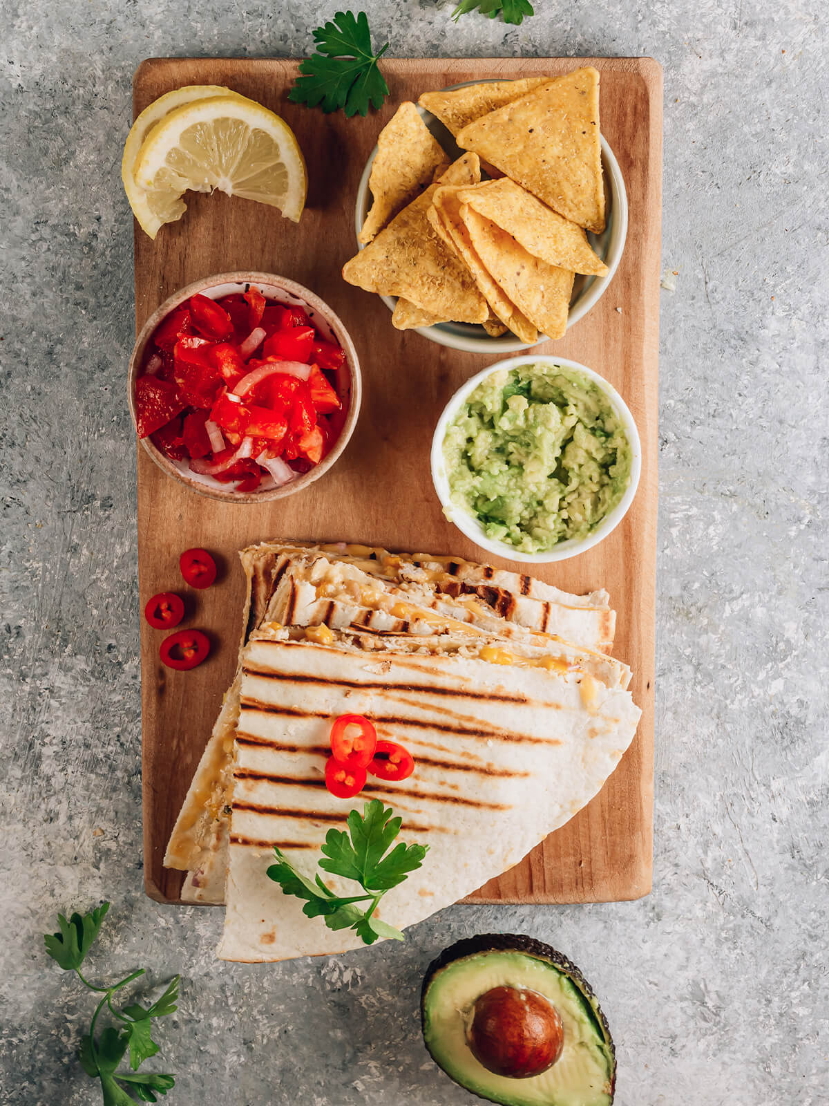 Gegrillte Quesadillas in Ecken geschnitten auf einem Holzbrett mit deutlichen Grillstreifen, dazu Tomaten-Salsa, Guacamole, Tortilla-Chips und eine Zitronenspalte