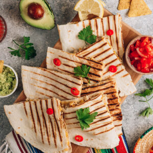 Gegrillte Quesadillas in Ecken geschnitten von oben auf einem Holzbrett, dazu Guacamole, Tomaten-Salsa, Tortilla-Chips und Zitrone auf bunter Decke