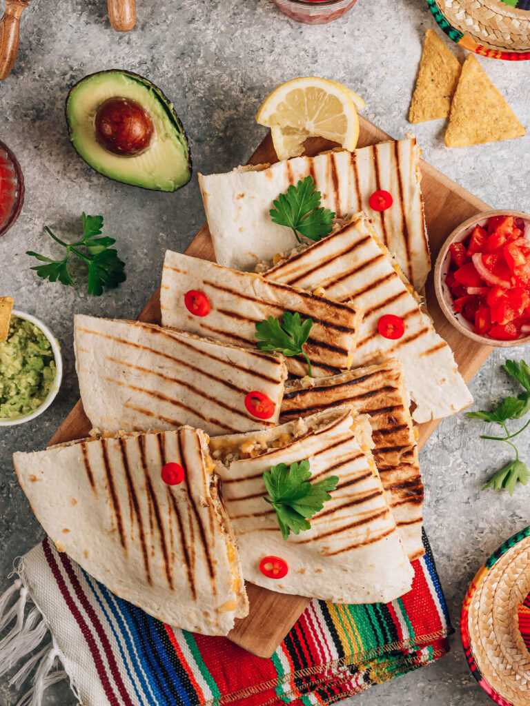 Gegrillte Quesadillas in Ecken geschnitten von oben auf einem Holzbrett, dazu Guacamole, Tomaten-Salsa, Tortilla-Chips und Zitrone auf bunter Decke
