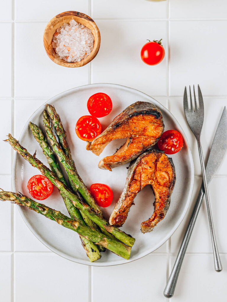 Gegrillter Lachs mit grünem Spargel, Kirschtomaten und grobem Salz auf hellem Teller, mediterran angerichtet von oben