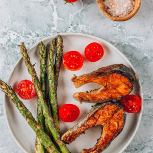 Gegrillter Lachs mit grünem Spargel, Kirschtomaten und grobem Salz auf einem hellen Teller, mediterran angerichtet