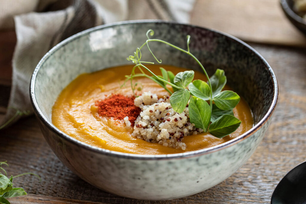 Geröstete Kürbis-Cremesuppe mit Räucherpaprika und Quinoa in einer hellen, grau-grün gesprenkelten Steingutschale, garniert mit dreifarbiger Quinoa, rotem Paprika-Staub und Erbsensprossen