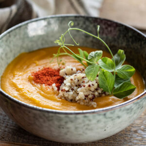 Geröstete Kürbis-Cremesuppe mit Räucherpaprika und Quinoa in einer hellen, grau-grün gesprenkelten Steingutschale, garniert mit dreifarbiger Quinoa, rotem Paprika-Staub und Erbsensprossen