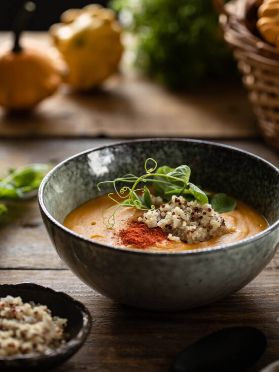 Geröstete Kürbis-Cremesuppe mit Räucherpaprika und Quinoa in heller, grau-grün gesprenkelter Steingutschale, garniert mit dreifarbiger Quinoa, rotem Paprika-Staub und Erbsensprossen