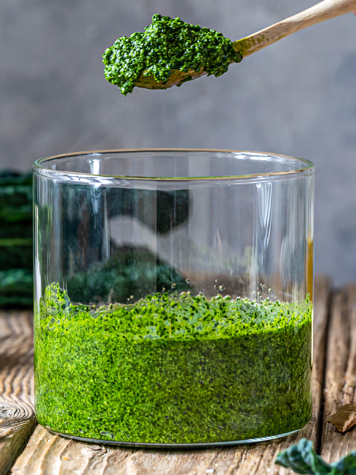 Grünkohl-Pesto mit Walnüssen, ein Holzlöffel mit tiefgrünem Pesto über einem Glas, dahinter frische Grünkohlblätter