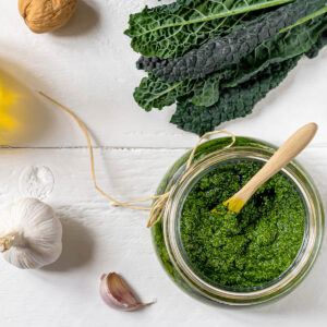 Grünkohl-Pesto mit Walnüssen in einem Glas, von oben fotografiert, umgeben von Grünkohlblättern, Knoblauch, Walnüssen, Parmesan und Olivenöl