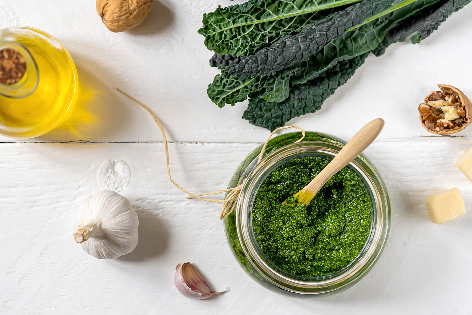 Grünkohl-Pesto von oben im Glas, umgeben von Grünkohlblättern, einer Knoblauchknolle, Walnüssen, einem Stück Parmesan und einer Flasche Olivenöl