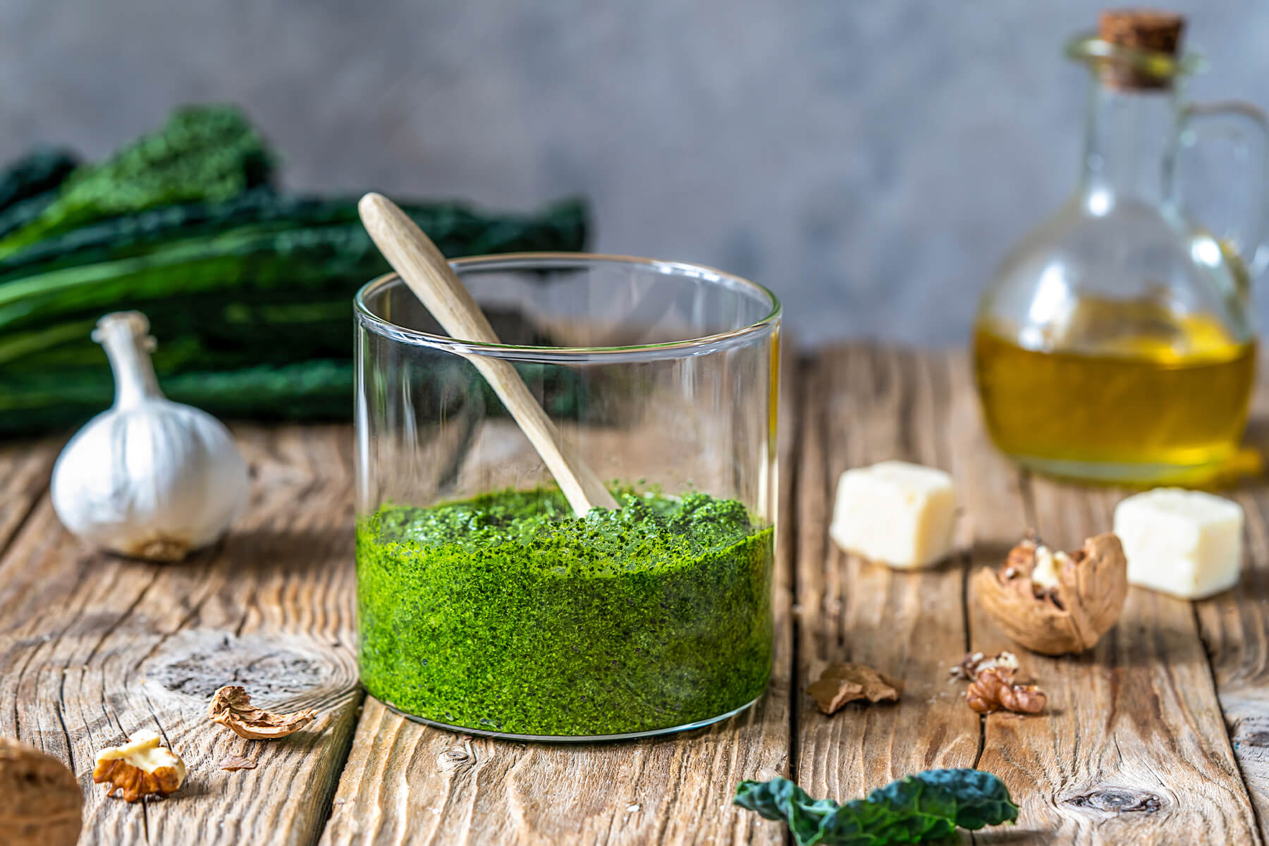 Frisch gemixtes Grünkohl-Pesto in einem Glas mit Holzlöffel, daneben eine Knoblauchknolle, Walnüsse, Parmesanwürfel und eine Karaffe Olivenöl