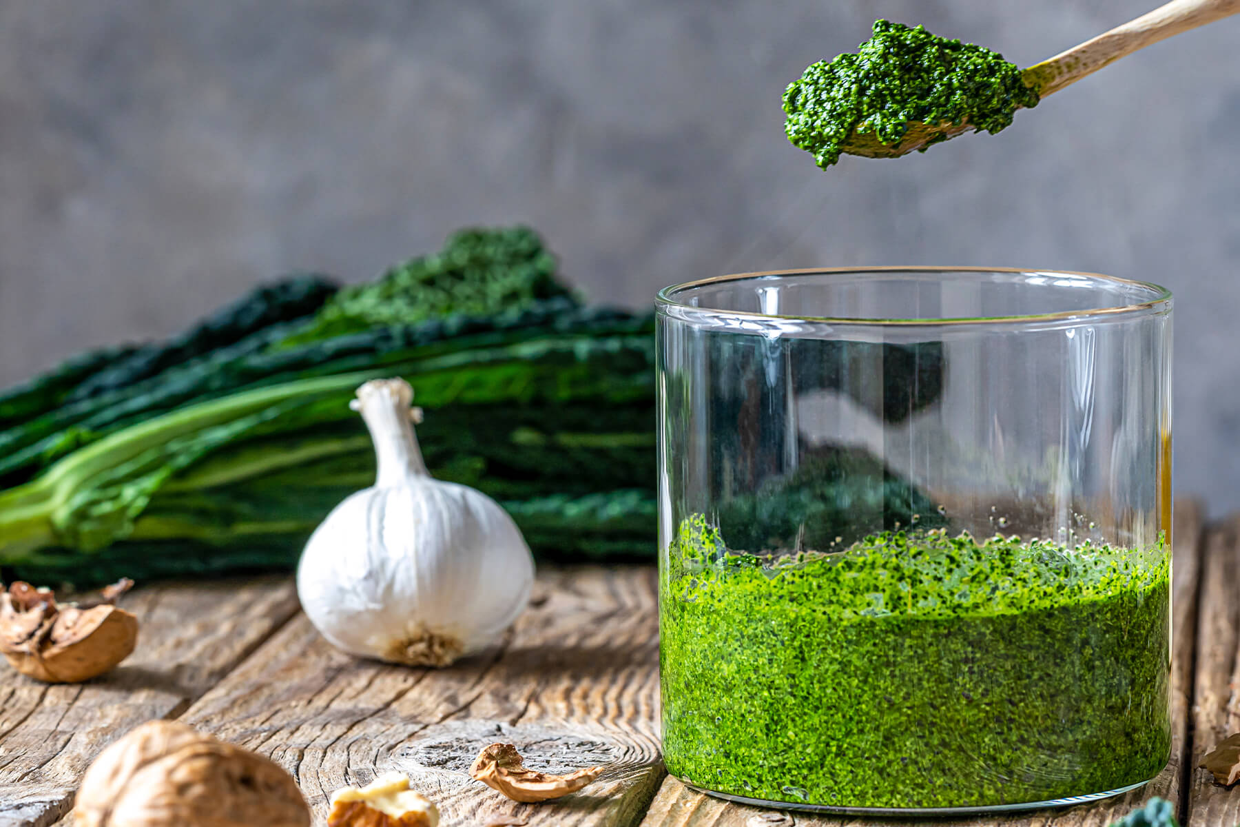 Ein Holzlöffel mit tiefgrünem Grünkohl-Pesto über einem Glas, dahinter frische Grünkohlblätter, eine Knoblauchknolle und eine Karaffe Olivenöl