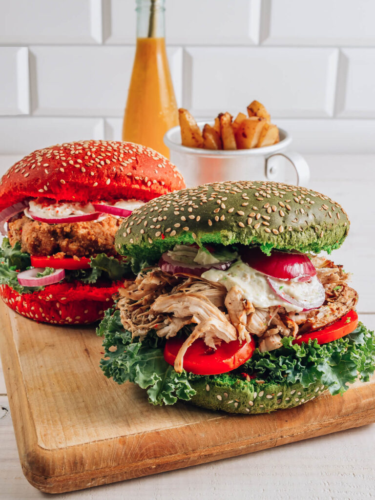 Zwei Hähnchen-Burger mit rotem und grünem Sesam-Brötchen, gefüllt mit knusprigem und gezupftem Hähnchen, Grünkohl, Tomate, roter Zwiebel und Kräutersauce