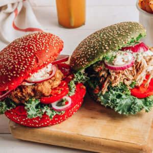 Zwei Hähnchen-Burger mit rotem und grünem Sesam-Brötchen auf einem Holzbrett, gefüllt mit Grünkohl, Tomate, roter Zwiebel und cremiger Kräutersauce