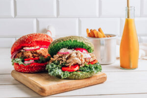 Fertig geschichteter roter und grüner Hähnchen-Burger auf einem Holzbrett, daneben Kartoffel-Spalten