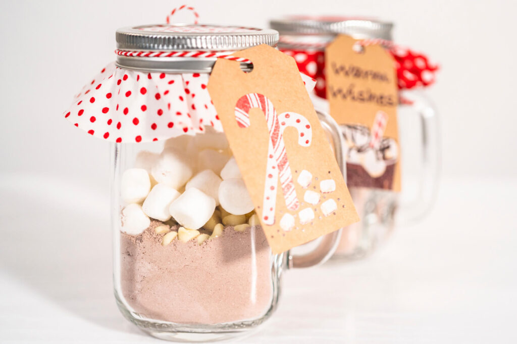 Weihnachtlich dekorierte Gläser mit geschichteter heißer Schokoladen-Mischung, hellen Schokoladen-Drops und Mini-Marshmallows, mit Punkte-Stoffdeckel, Bäckergarn und Geschenkanhänger