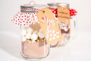 Weihnachtlich dekorierte Gläser mit geschichteter heißer Schokoladen-Mischung, hellen Schokoladen-Drops und Mini-Marshmallows, mit Punkte-Stoffdeckel, Bäckergarn und Geschenkanhänger