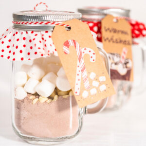 Weihnachtlich dekorierte Gläser mit geschichteter heißer Schokoladen-Mischung, hellen Schokoladen-Drops und Mini-Marshmallows, mit Punkte-Stoffdeckel, Bäckergarn und Geschenkanhänger