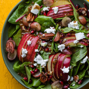 Herbstsalat mit gefächerten Apfelhälften, Trauben, Granatapfel und Ziegenkäse von oben in einer grünen Schale vor leuchtend gelbem Hintergrund
