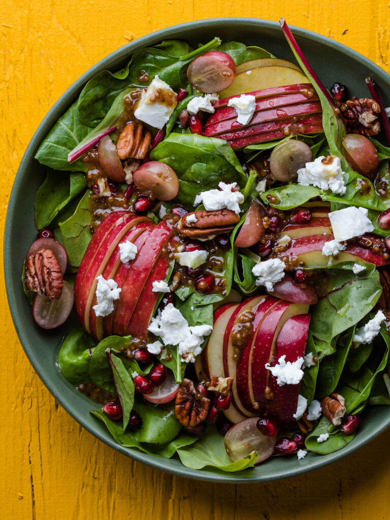 Herbstsalat mit gefächerten Apfelhälften, Trauben, Granatapfel und Ziegenkäse von oben in einer grünen Schale vor leuchtend gelbem Hintergrund