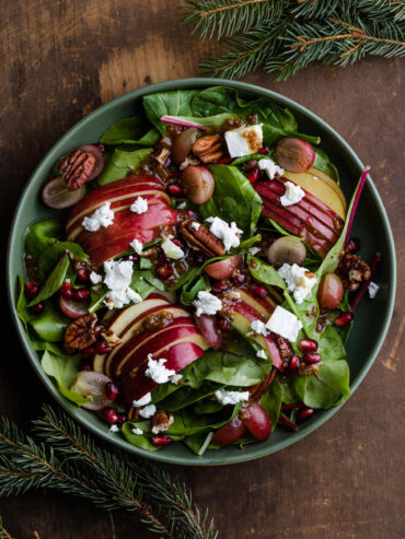 Herbstsalat mit gefächerten Apfelhälften, roten und hellen Weintrauben, Pekannüssen, Ziegenkäse und Granatapfelkernen in einer grünen Schale, von oben fotografiert mit Tannenzweigen