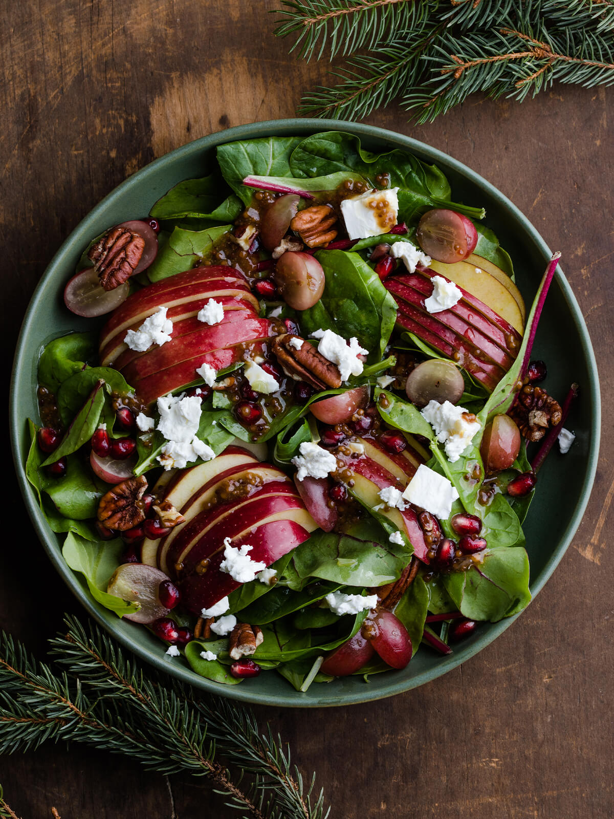 Herbstsalat mit gefächerten Apfelhälften, roten und hellen Weintrauben, Pekannüssen, Ziegenkäse und Granatapfelkernen in einer grünen Schale, von oben fotografiert mit Tannenzweigen