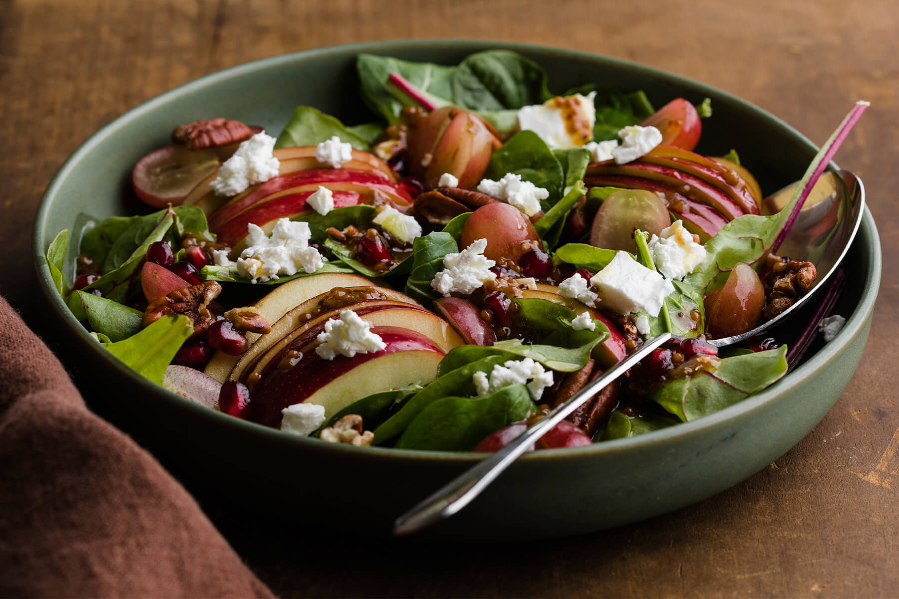 Nahaufnahme des Herbstsalats mit gefächerten Apfelscheiben, halbierten Trauben, Pekannüssen, Ziegenkäse-Bröckchen und Granatapfelkernen, beträufelt mit einer Vinaigrette mit sichtbaren Senfkörnern
