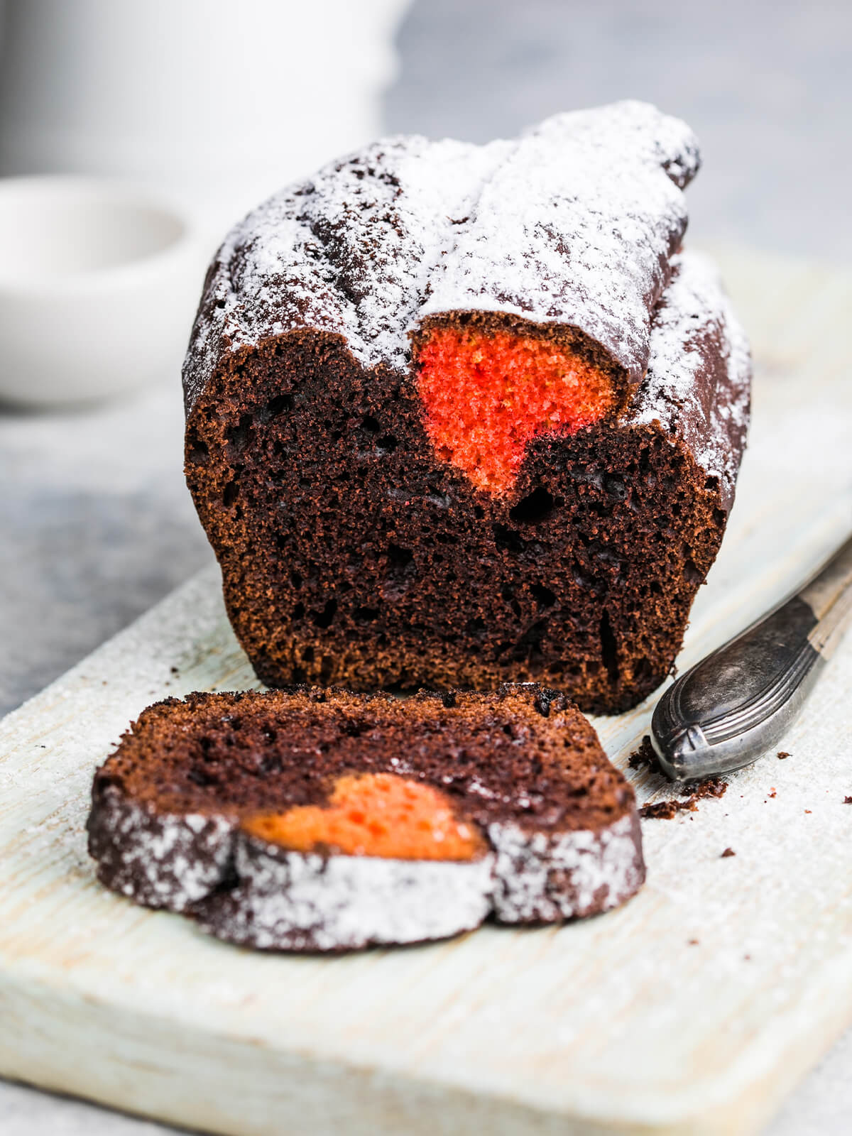 Angeschnittener dunkler Schoko-Kastenkuchen mit Puderzucker, in dessen Schnittfläche ein leuchtend orange-rotes Herz erscheint, davor drei abgeschnittene Scheiben