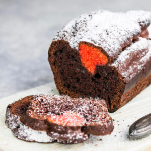 Schoko-Kastenkuchen mit Puderzucker auf einem Holzbrett mit Messer, davor drei abgeschnittene Scheiben, in der vordersten leuchtet das orange-rote Herz