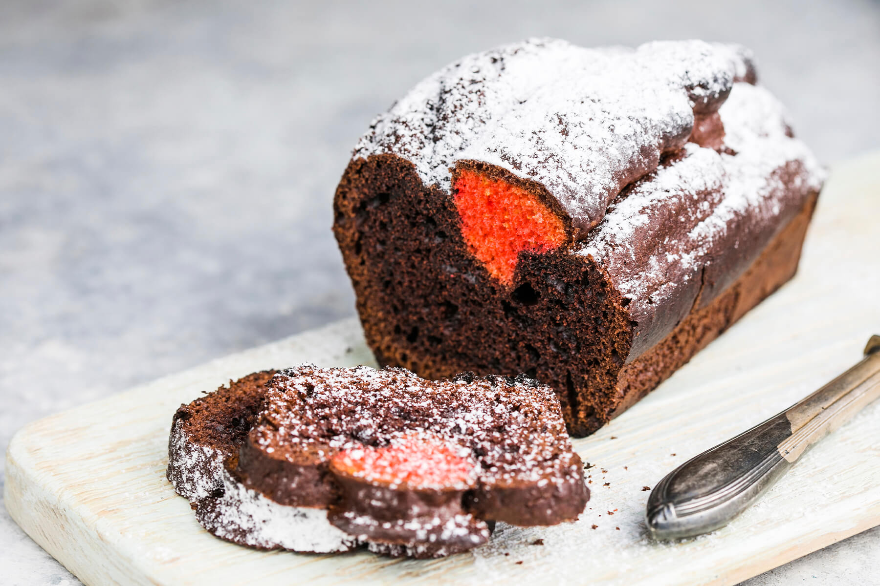 Schoko-Kastenkuchen mit Puderzucker auf einem Holzbrett mit Messer, davor drei abgeschnittene Scheiben, in der vordersten leuchtet das orange-rote Herz