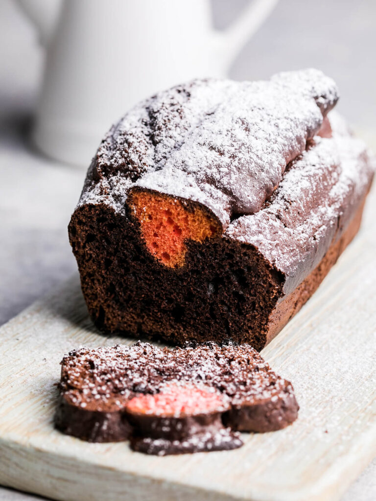 Dunkler Schoko-Kastenkuchen mit Puderzucker auf einem Holzbrett, in der Schnittfläche ein orange-rotes Herz, davor drei abgeschnittene Scheiben, im Hintergrund eine weiße Kanne
