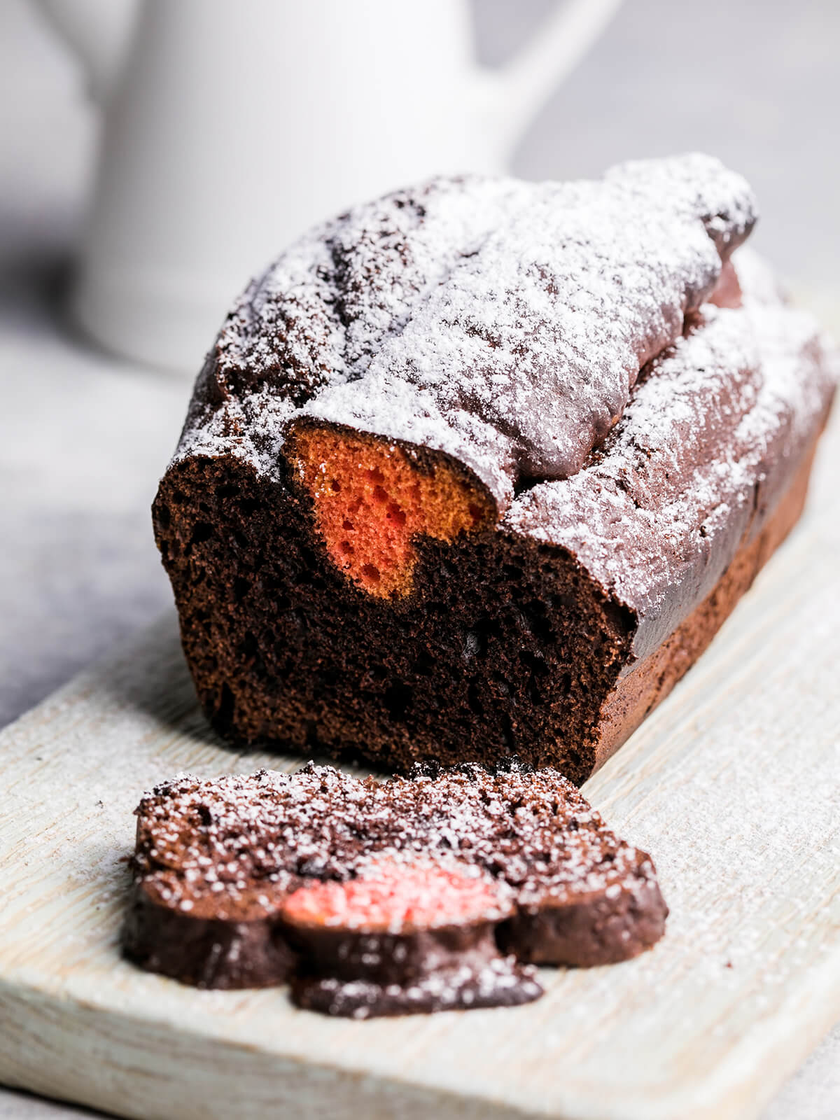 Dunkler Schoko-Kastenkuchen mit Puderzucker auf einem Holzbrett, in der Schnittfläche ein orange-rotes Herz, davor drei abgeschnittene Scheiben, im Hintergrund eine weiße Kanne