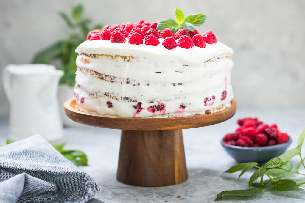Himbeer-Sahne-Torte als Naked Cake von der Seite, vier Biskuitböden mit weisser Sahnecreme und Himbeeren zwischen den Schichten auf einem Holz-Tortenständer