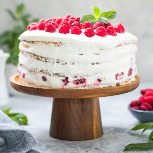Himbeer-Sahne-Torte als Naked Cake von der Seite, vier Biskuitböden mit weisser Sahnecreme und Himbeeren zwischen den Schichten auf einem Holz-Tortenständer