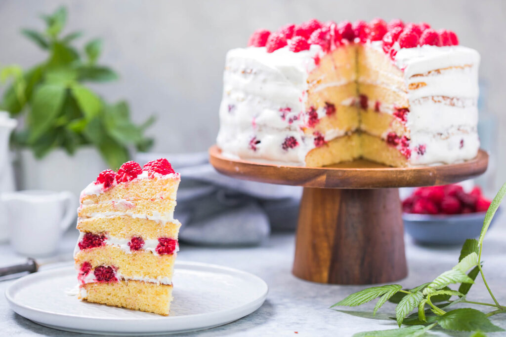 Angeschnittene Himbeer-Sahne-Torte, ein Stück auf einem Teller zeigt vier hellgelbe Biskuitböden mit weißer Sahnecreme und ganzen Himbeeren in den drei Füllschichten