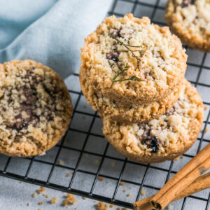 Runde Streusel-Plätzchen mit Himbeermarmelade auf einem schwarzen Kuchengitter, daneben Zimtstangen und kleine getrocknete Blüten