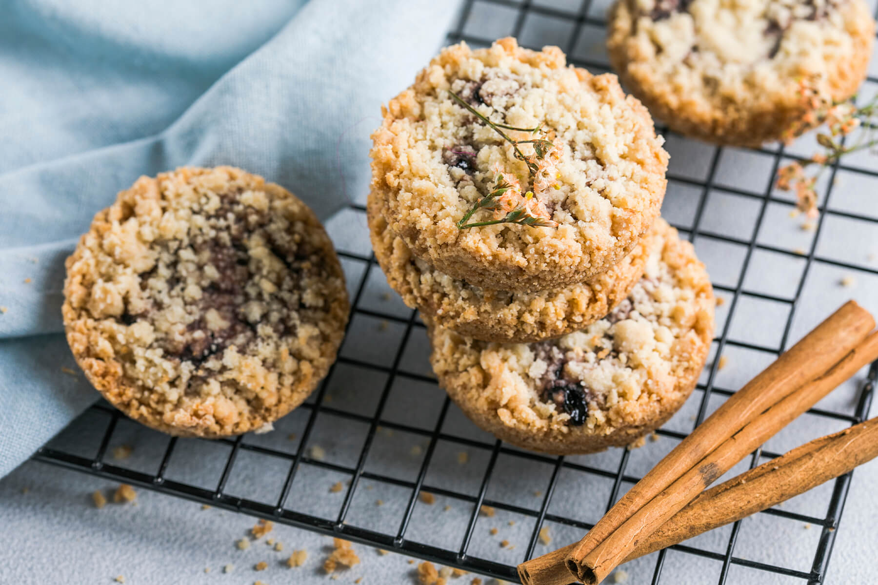 Runde Streusel-Plätzchen mit Himbeermarmelade auf einem schwarzen Kuchengitter, daneben Zimtstangen und ein hellblaues Tuch