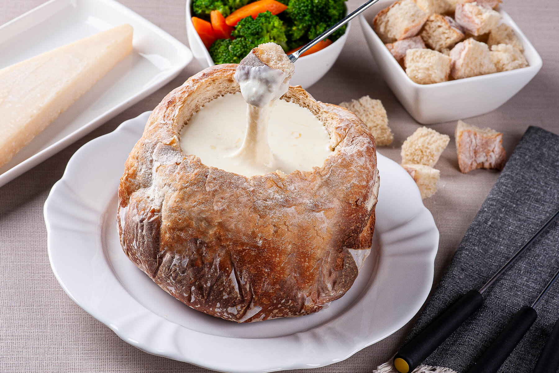 Fonduegabel hebt ein Stück Brot mit geschmolzenem Käse aus einem ausgehöhlten Brotlaib, daneben Schälchen mit Brokkoli, Karotte und Brotwürfeln