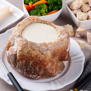 Rundes ausgehöhltes Landbrot gefüllt mit cremig geschmolzenem Käse, daneben Fonduegabeln und Schälchen mit Brokkoli, Karotte und Brotwürfeln