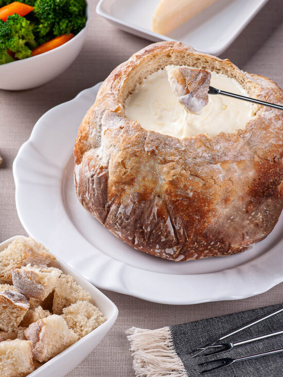 Rundes rustikales Landbrot als essbare Schale, gefüllt mit cremig geschmolzenem Käsefondue, eine Fonduegabel taucht ein