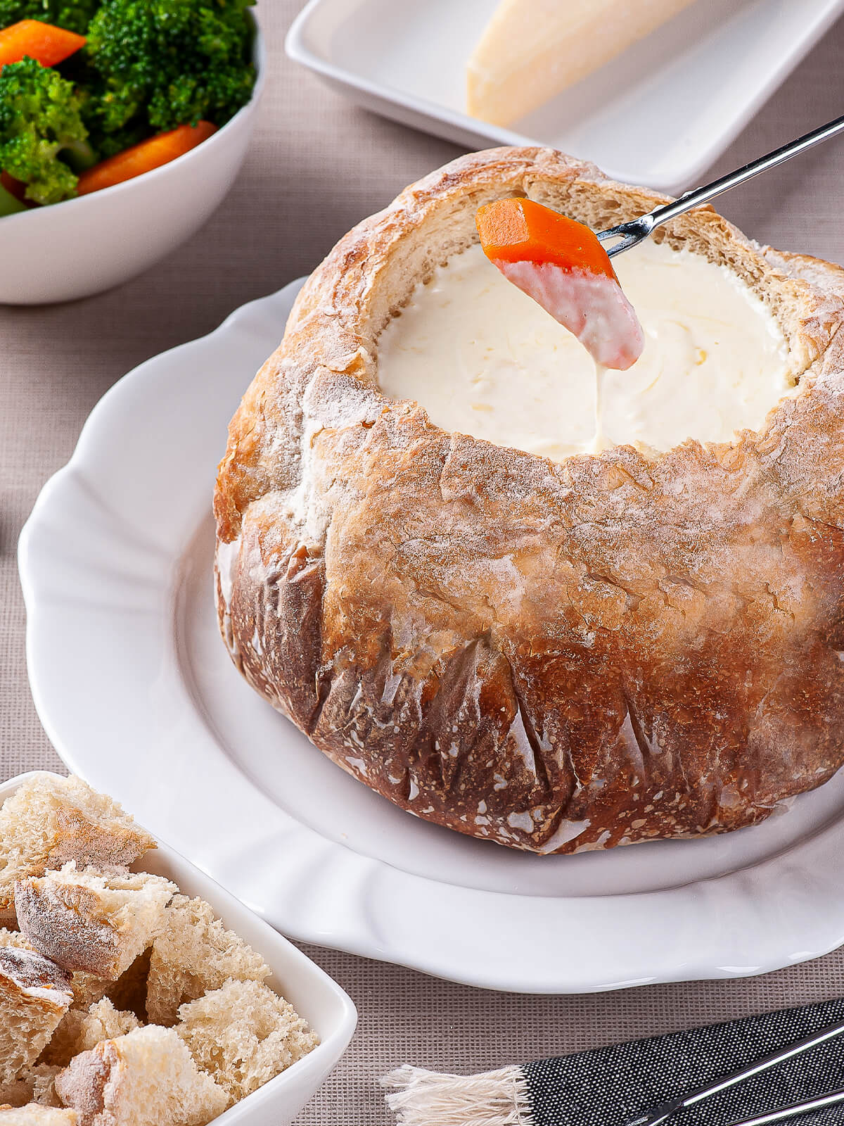 Fonduegabel taucht ein Stück Karotte in das cremig geschmolzene Käsefondue im Brotlaib, daneben eine Schale mit Brokkoli und Karotte