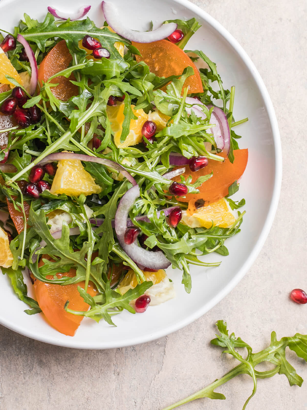 Kaki-Rucola-Salat mit Orangenfilets, roten Zwiebelringen und Granatapfelkernen auf einem weißen Teller von oben
