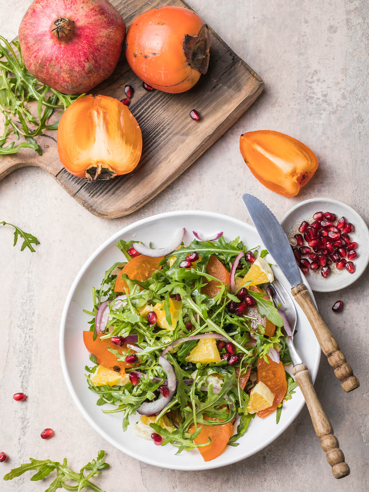 Kaki-Rucola-Salat in weißer Schale neben einem Holzbrett mit ganzem Granatapfel, ganzen und aufgeschnittenen Kakis sowie einem Schälchen Granatapfelkerne