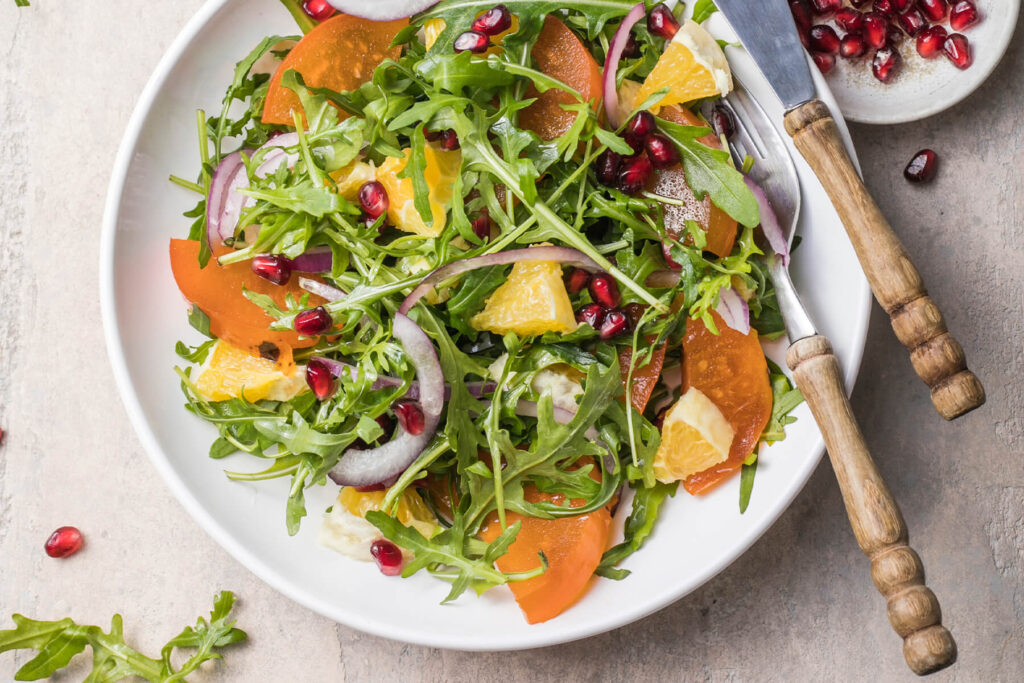 Kaki-Rucola-Salat mit Orangenfilets, roten Zwiebelringen und Granatapfelkernen in einer weißen Schale von oben