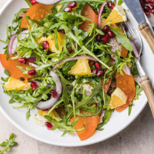 Kaki-Rucola-Salat mit Orangenfilets, roten Zwiebelringen und Granatapfelkernen in einer weißen Schale von oben