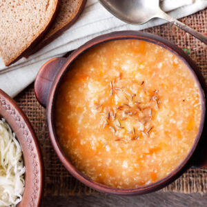 Dicke orange-braune Sauerkrautsuppe mit Hirse und Kümmel in einer Tonschale, daneben Roggenbrot und eine kleine Schale rohes Sauerkraut