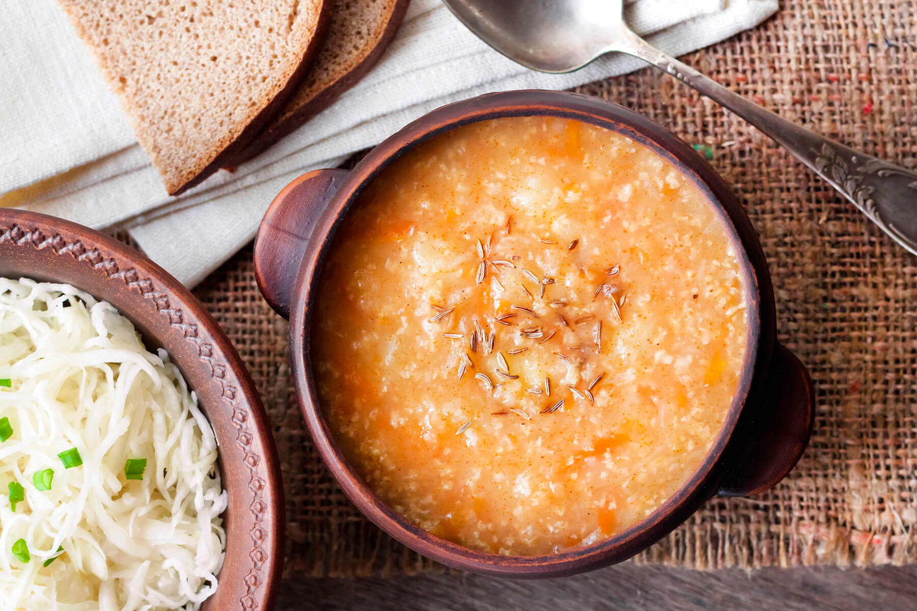 Draufsicht auf eine Tonschale mit dicker Sauerkrautsuppe, Hirse und Kümmel, daneben Roggenbrot und eine kleine Schale rohes Sauerkraut