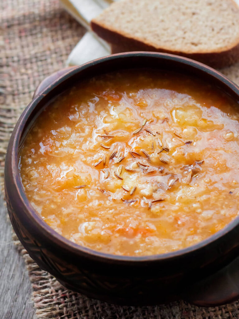 Nahaufnahme einer Tonschale mit sämiger orange-brauner Sauerkrautsuppe, obenauf deutlich gestreuter Kümmel, im Hintergrund Roggenbrot