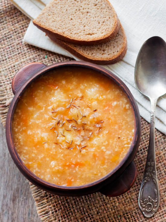 Dicke orange-braune ukrainische Sauerkrautsuppe mit Hirse und Kümmel in einer Tonschale, daneben zwei Scheiben dunkles Roggenbrot und ein Löffel